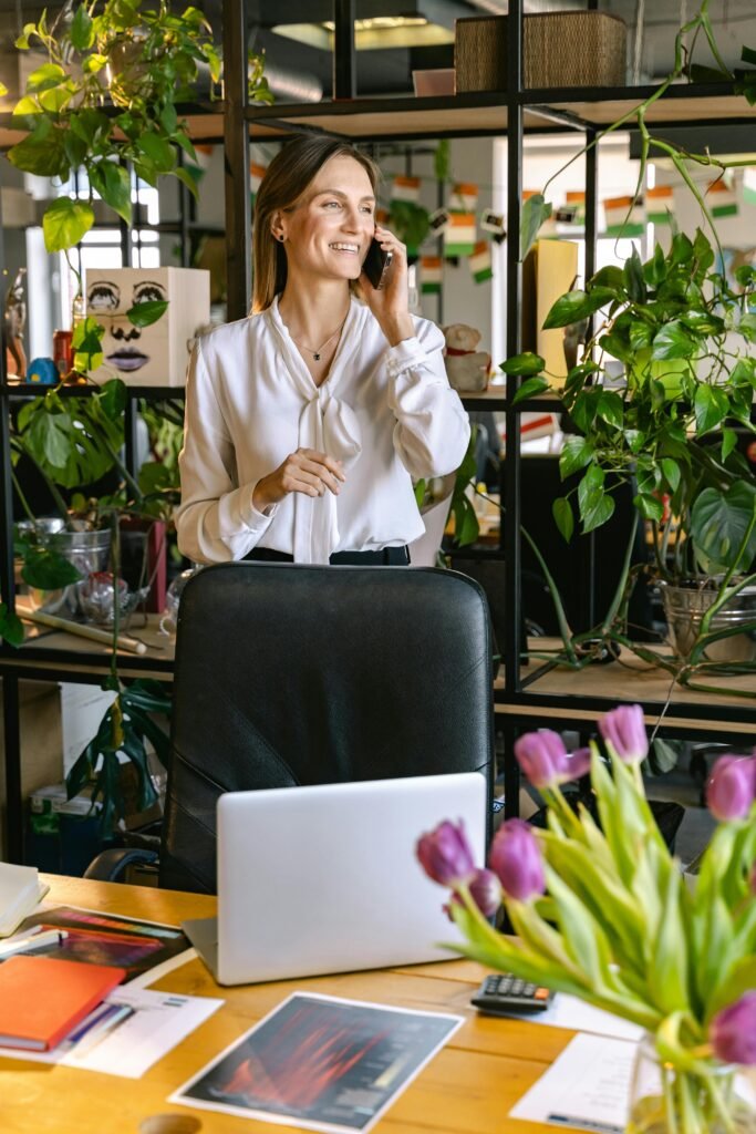 pexels photo 7163400 7163400 A woman smiling and talking on a phone in a bright, plant-filled office setting, standing by her desk.